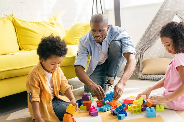 Padre afroamericano y los niños jugando con bloques de colores en el piso juntos en casa - foto de stock