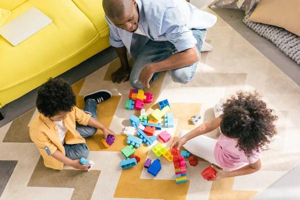 Vue aérienne du père afro-américain et des enfants jouant avec des blocs colorés sur le sol ensemble à la maison — Photo de stock