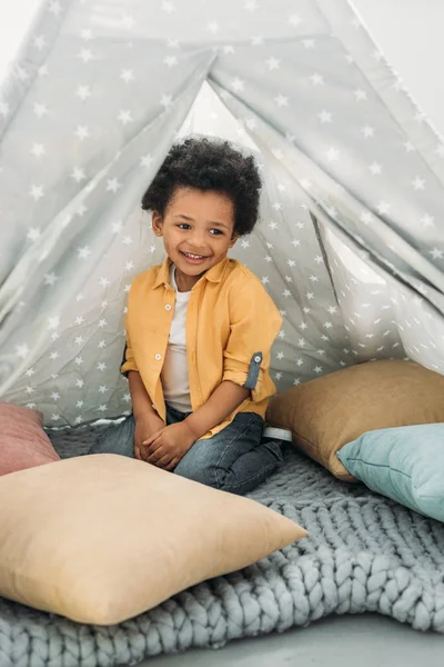 Petit afro-américain souriant assis dans un tipi à la maison — Photo de stock