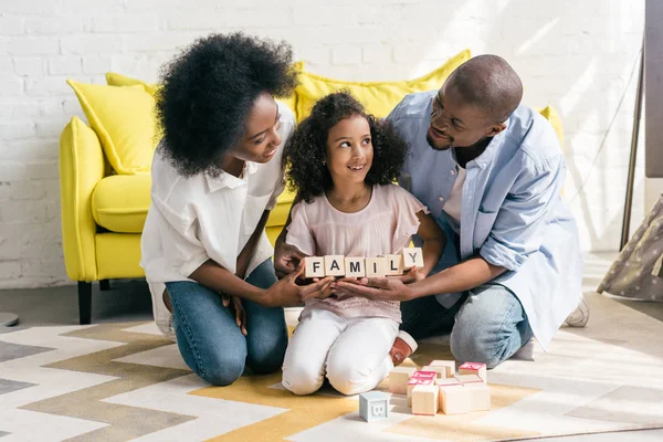 Африканские американские родители и дочь держат деревянные блоки с семейными надписями вместе на полу дома — стоковое фото