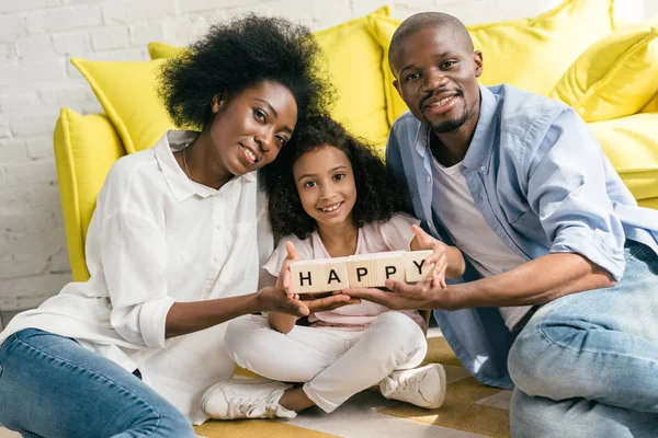 African american parents and daughter holding wooden blocks with happy lettering together on floor at home — Stock Photo