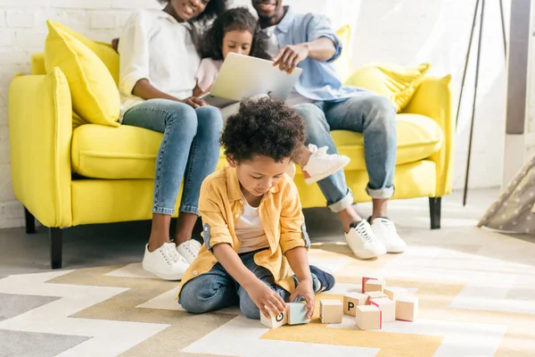 Enfoque selectivo de niño afroamericano jugando con bloques de madera, mientras que los padres y la hermana usando el ordenador portátil en el sofá en casa - foto de stock