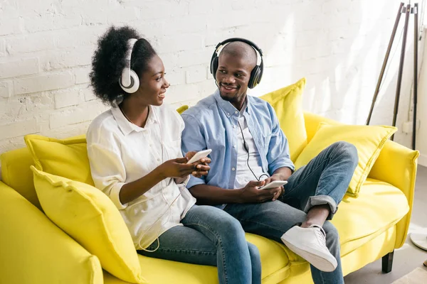 Afroamericanos escuchando música en auriculares con teléfonos inteligentes mientras descansan en el sofá en casa - foto de stock