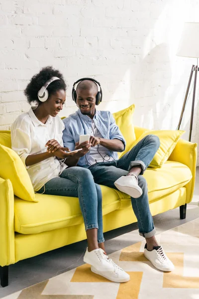 Afro-americano ouvir música em fones de ouvido e usando smartphones enquanto descansa no sofá em casa — Fotografia de Stock