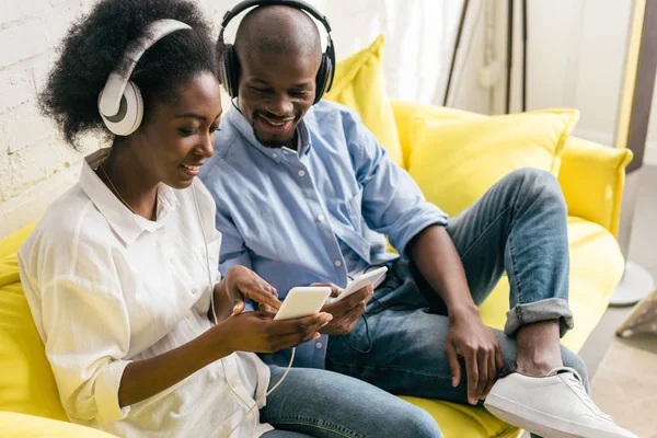 Sonriente afroamericano escuchando música en auriculares y usando teléfonos inteligentes mientras descansa en el sofá en casa - foto de stock