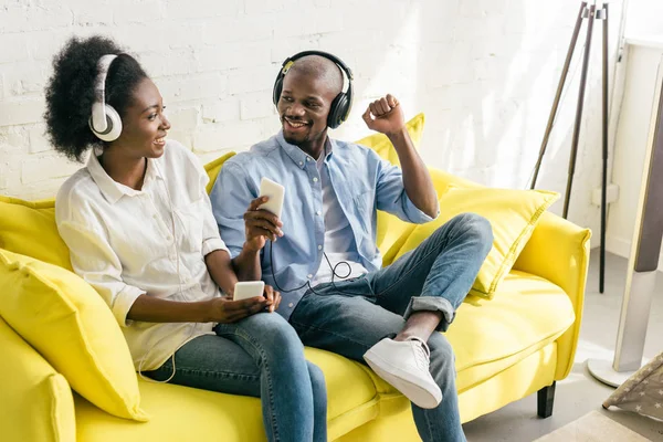 Alegre afro-americano ouvir música em fones de ouvido com smartphones enquanto descansa no sofá em casa — Fotografia de Stock