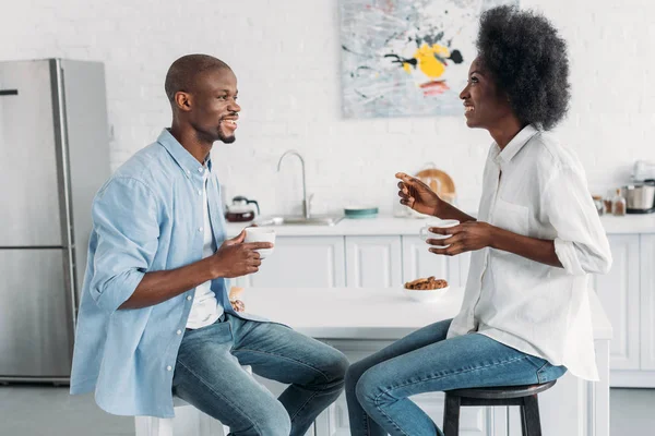 Vista lateral de pareja afroamericana con tazas de café en la mañana juntos en la cocina en casa - foto de stock