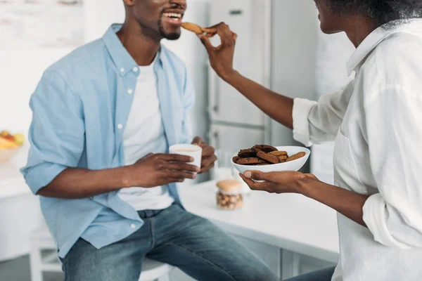 Partial view of african american woman feeding husband with cookies in morning in kitchen at home — Stock Photo