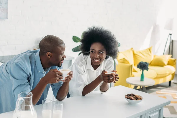 Sourire couple afro-américain avec des tasses de café le matin ensemble dans la cuisine à la maison — Photo de stock