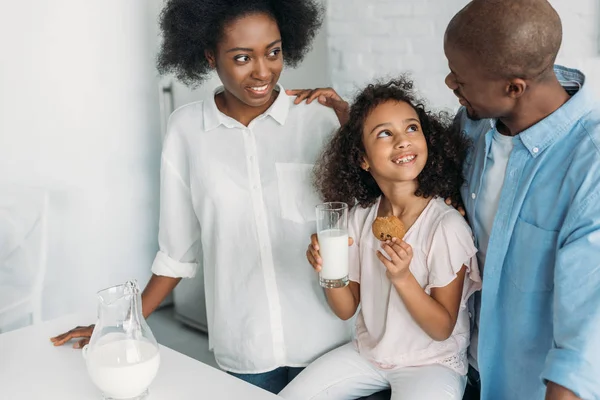 Bambino afroamericano sorridente con bicchiere di latte, biscotti in mano e genitori vicini in cucina a casa — Foto stock