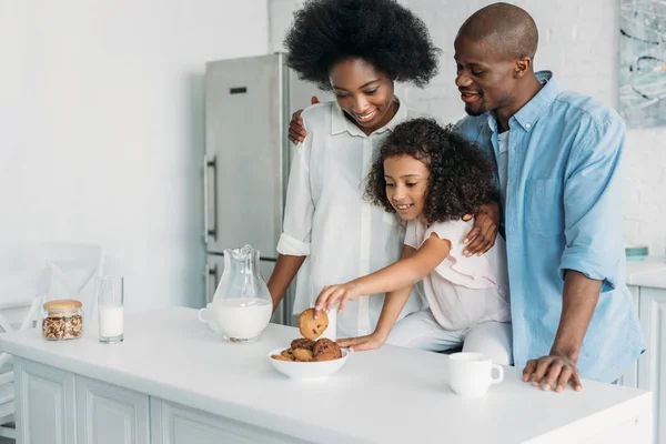 Criança americana africana tomando biscoito enquanto os pais estão perto na cozinha em casa — Fotografia de Stock