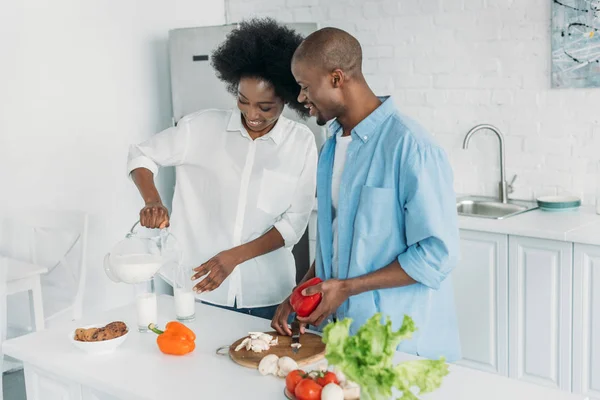 Улыбающаяся африканская американка наливает молоко в стакан, а муж готовит завтрак на кухне дома — стоковое фото