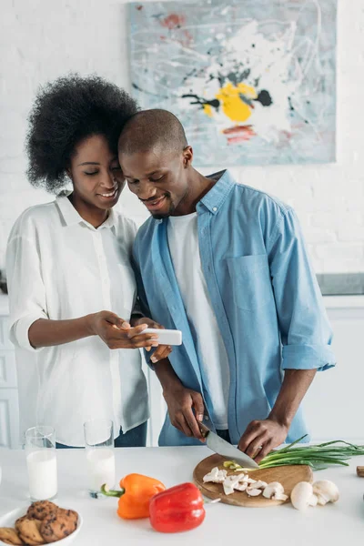 Улыбающийся африканский американец готовит завтрак и использует смартфон вместе с женой на кухне дома — стоковое фото