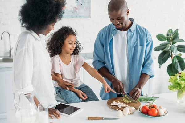 Hombre afroamericano cortando verduras frescas para el desayuno con la familia de pie cerca en la cocina en casa - foto de stock