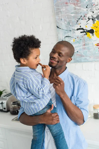 Uomo afroamericano che tiene in braccio il piccolo figlio con biscotto in cucina a casa — Foto stock