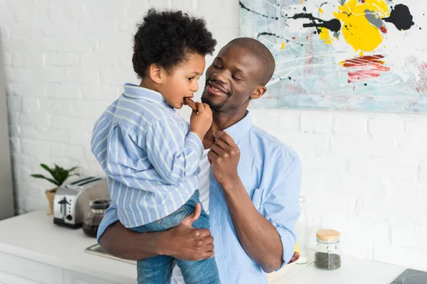 African american man holding little son with cookie in kitchen at home — Stock Photo