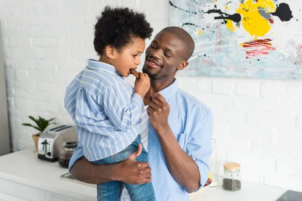 Afro-americano homem segurando pequeno filho com biscoito na cozinha em casa — Fotografia de Stock