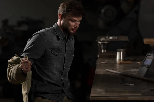 Junger männlicher Arbeiter zieht Uniform aus und sitzt in der Fabrik am Tisch — Stockfoto