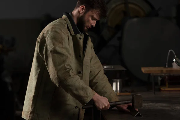 Visão lateral do jovem trabalhador com martelo e peça de metal na fábrica — Fotografia de Stock