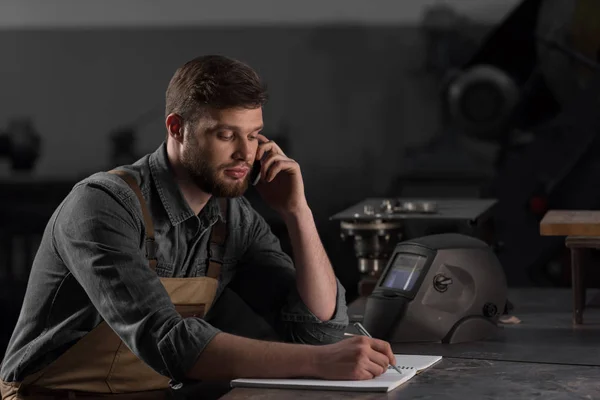 Männliche Arbeiter schreiben in Lehrbuch und sprechen auf dem Smartphone — Stockfoto