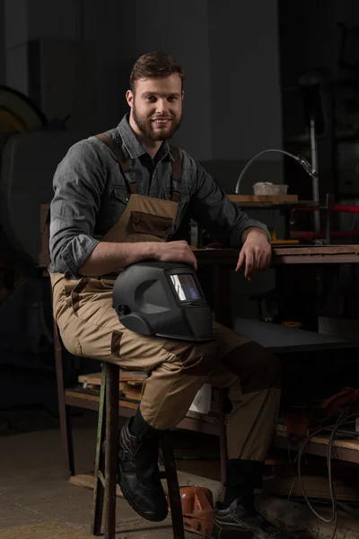 Trabalhador sorrindo em uniforme segurando máscara protetora na fábrica — Fotografia de Stock