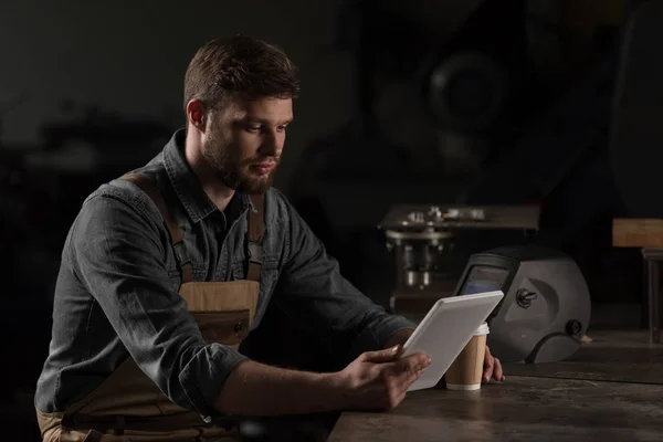 Jeune ouvrier masculin avec tasse en papier de café en utilisant une tablette numérique — Photo de stock