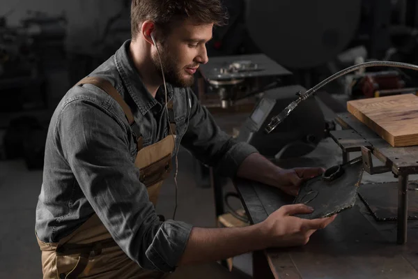 Joven trabajador masculino en auriculares escuchando música y mirando parte del metal - foto de stock