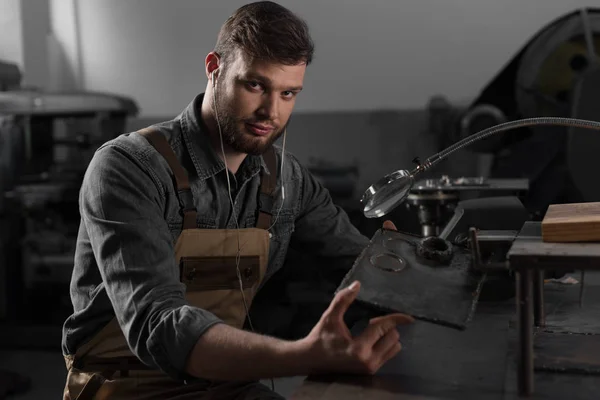 Portrait of young male worker in earphones holding metal part under magnifying glass — Stock Photo