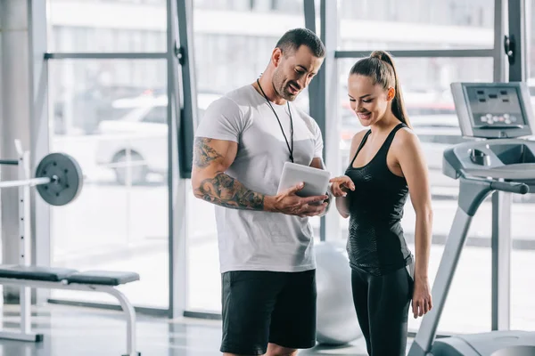 Sorridente jovem esportista apontando por dedo no cronograma em tablet digital em mãos personal trainer no ginásio — Fotografia de Stock