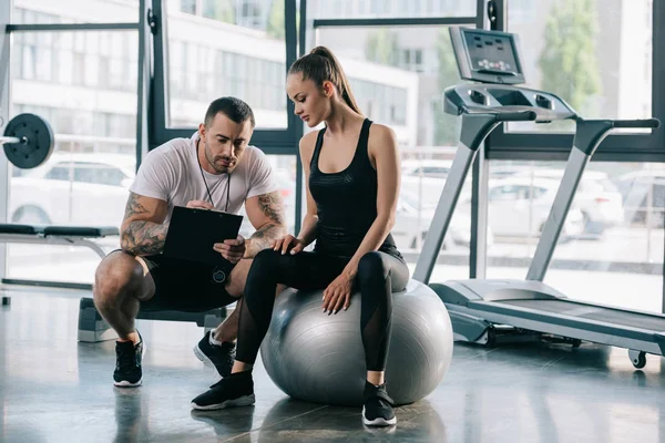 Treinador masculino escrevendo em prancheta e jovem esportista sentado na bola de fitness no ginásio — Fotografia de Stock