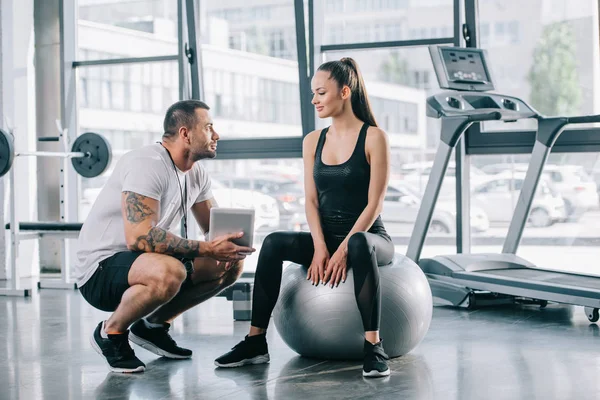 Giovane sportiva seduta sulla palla fitness e che parla con personal trainer con tablet digitale in palestra — Foto stock