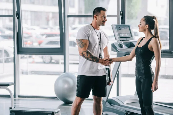 Smiling male personal trainer and young sportswoman shaking hands at gym — Stock Photo