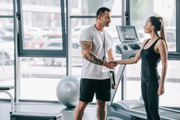 Sonriente entrenador personal masculino y joven deportista estrechando la mano en el gimnasio - foto de stock