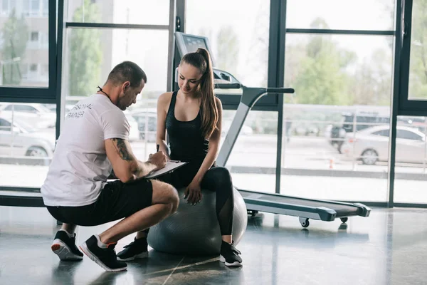 Männliche Trainerin schreibt in Klemmbrett und junge Sportlerin sitzt im Fitnessstudio auf Fitnessball — Stockfoto