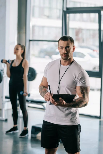 Personal trainer com prancheta e jovem desportista exercitando com halteres atrás no ginásio — Fotografia de Stock