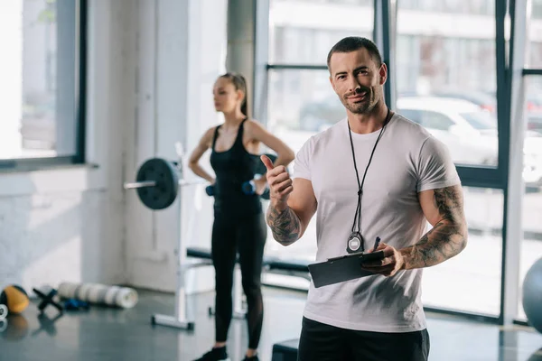 Personal trainer with clipboard doing thumb up gesture and young sportswoman exercising with dumbbells behind at gym — Stock Photo