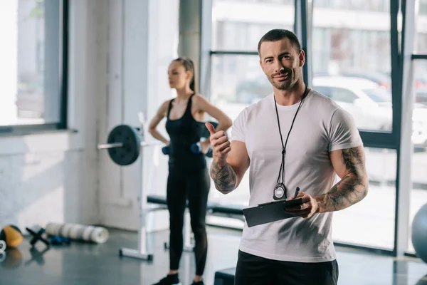 Entrenador personal con portapapeles haciendo gesto de pulgar hacia arriba y joven deportista haciendo ejercicio con pesas detrás en el gimnasio - foto de stock