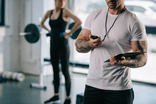 Cropped shot of male personal trainer looking at timer and young sportswoman exercising with dumbbells behind at gym — Stock Photo