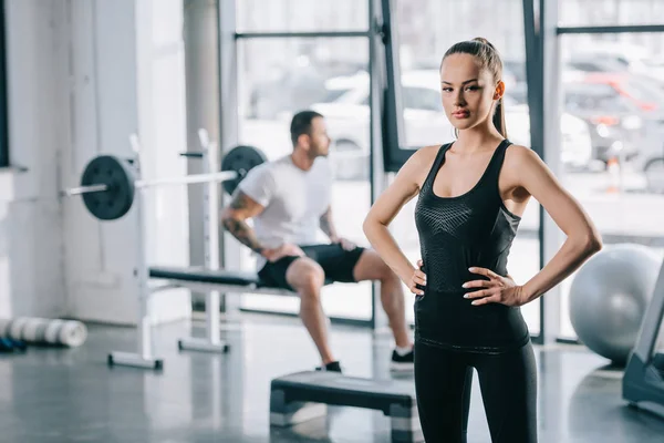 Young sportswoman with hands on thighs and man sitting behind at gym — Stock Photo