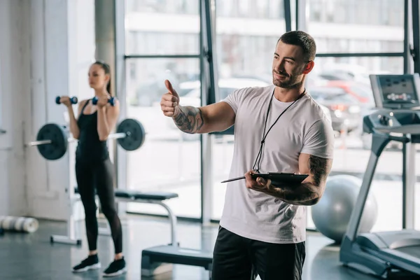 Entrenador personal haciendo gesto pulgar hacia arriba y joven deportista haciendo ejercicio con mancuernas detrás en el gimnasio - foto de stock