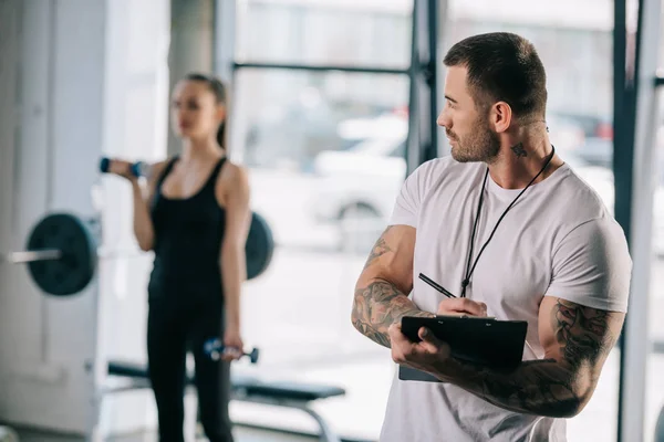 Entrenador personal masculino escribiendo en portapapeles y joven atlética haciendo ejercicio con pesas detrás en el gimnasio - foto de stock