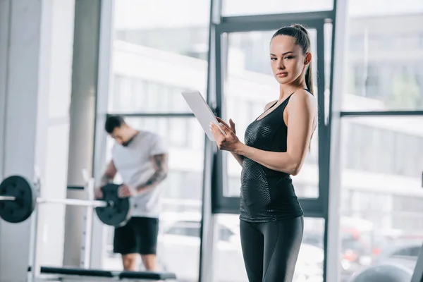 Young female trainer with digital tablet and athletic man with barbell at gym — Stock Photo