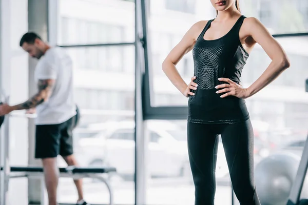 Tiro recortado de mujer con las manos en los muslos y el hombre atlético con la barra detrás en el gimnasio - foto de stock