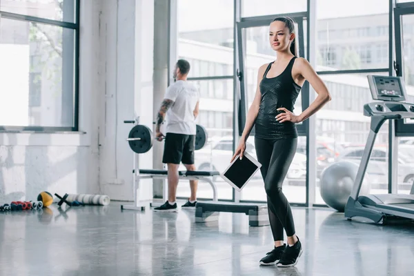 Jovem personal trainer feminino com tablet digital e homem atlético com barra atrás no ginásio — Fotografia de Stock