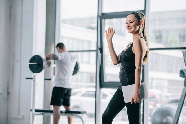 Personaltrainerin mit digitalem Tablet, winkende Hand und athletischer Mann, der mit Langhantel im Rücken im Fitnessstudio trainiert — Stockfoto