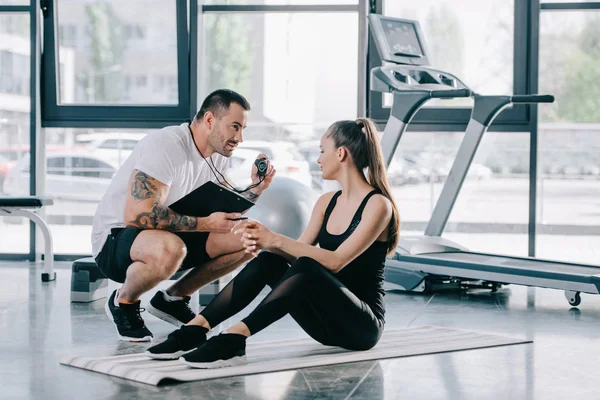 Male personal trainer showing timer to young athletic woman on fitness mat at gym — Stock Photo