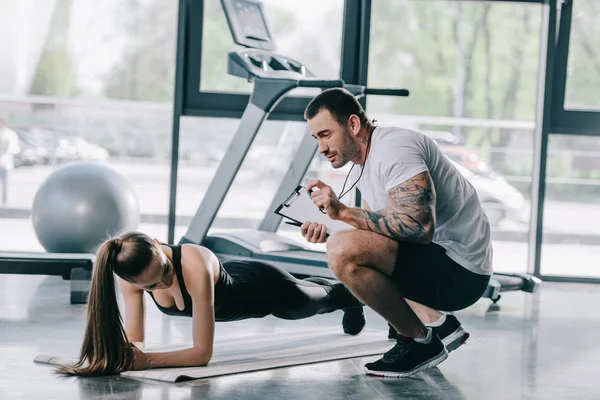 Male personal trainer showing timer to young athletic woman doing plank on fitness mat at gym — Stock Photo