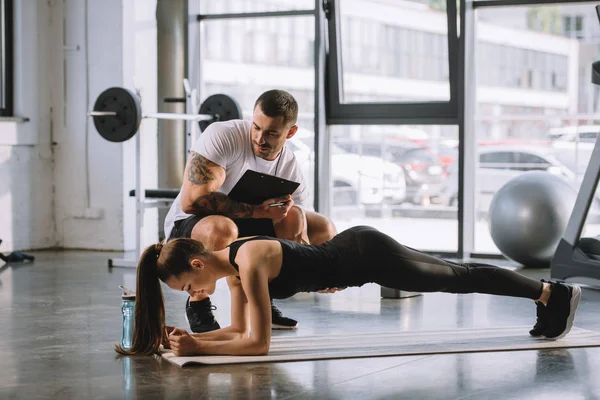 Male personal trainer with clipboard and young athletic woman doing plank on fitness mat at gym — Stock Photo