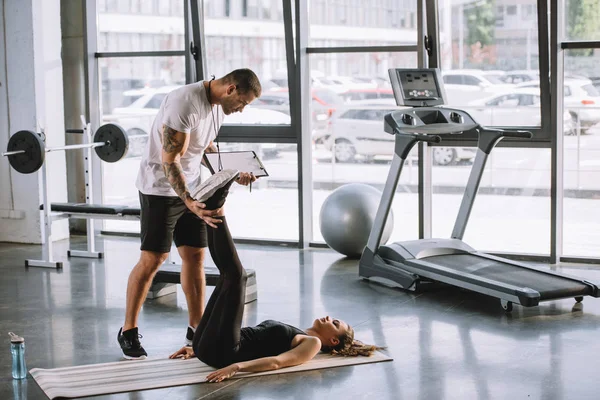 Masculino personal trainer ajudando jovem atlético mulher para fazer abs no tapete de fitness no ginásio — Fotografia de Stock