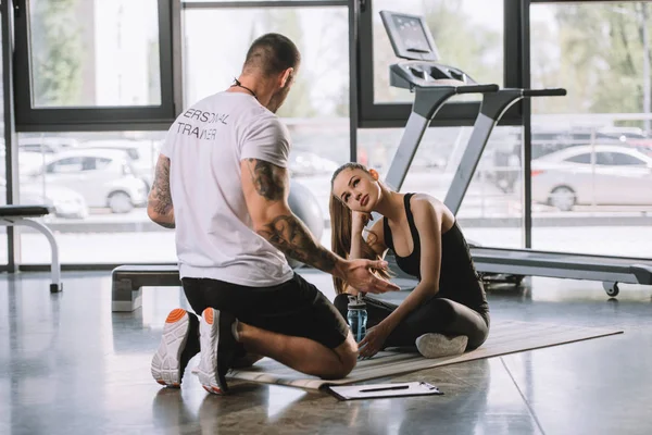 Entrenador personal masculino explicando hablar con una joven atlética sentada en una colchoneta de fitness en el gimnasio - foto de stock